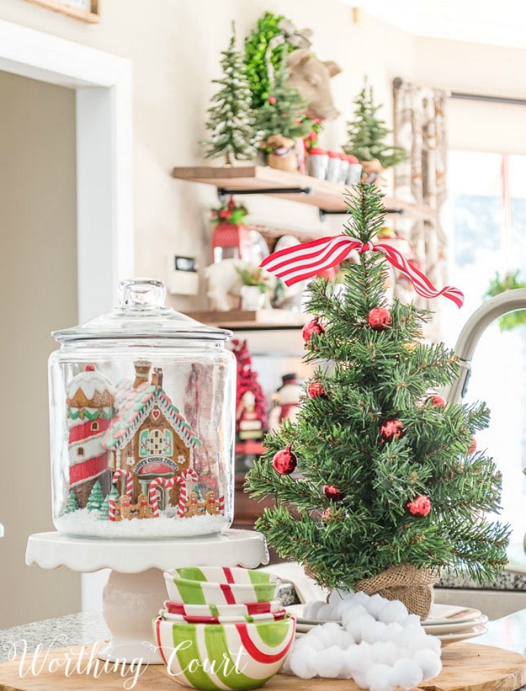 A glass jar with gingerbread house inside it on a cake stand beside a tabletop Christmas tree.