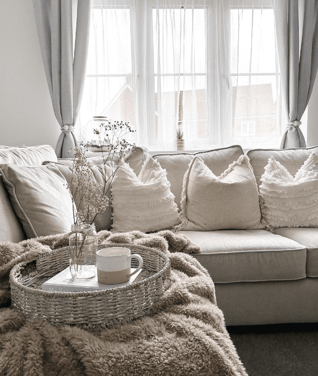 Living room featuring neutral and light blue sofa with pillows and a large window in the background with a combination of window treatments