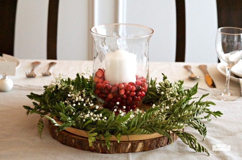Centerpiece idea featuring large white pillar candle with greenery, red berries on top of a wood slice