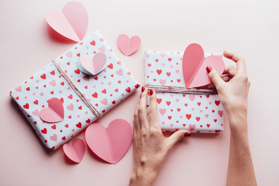 Valentine's Day stock photo featuring hands wrapping gift with pink background and paper hearts