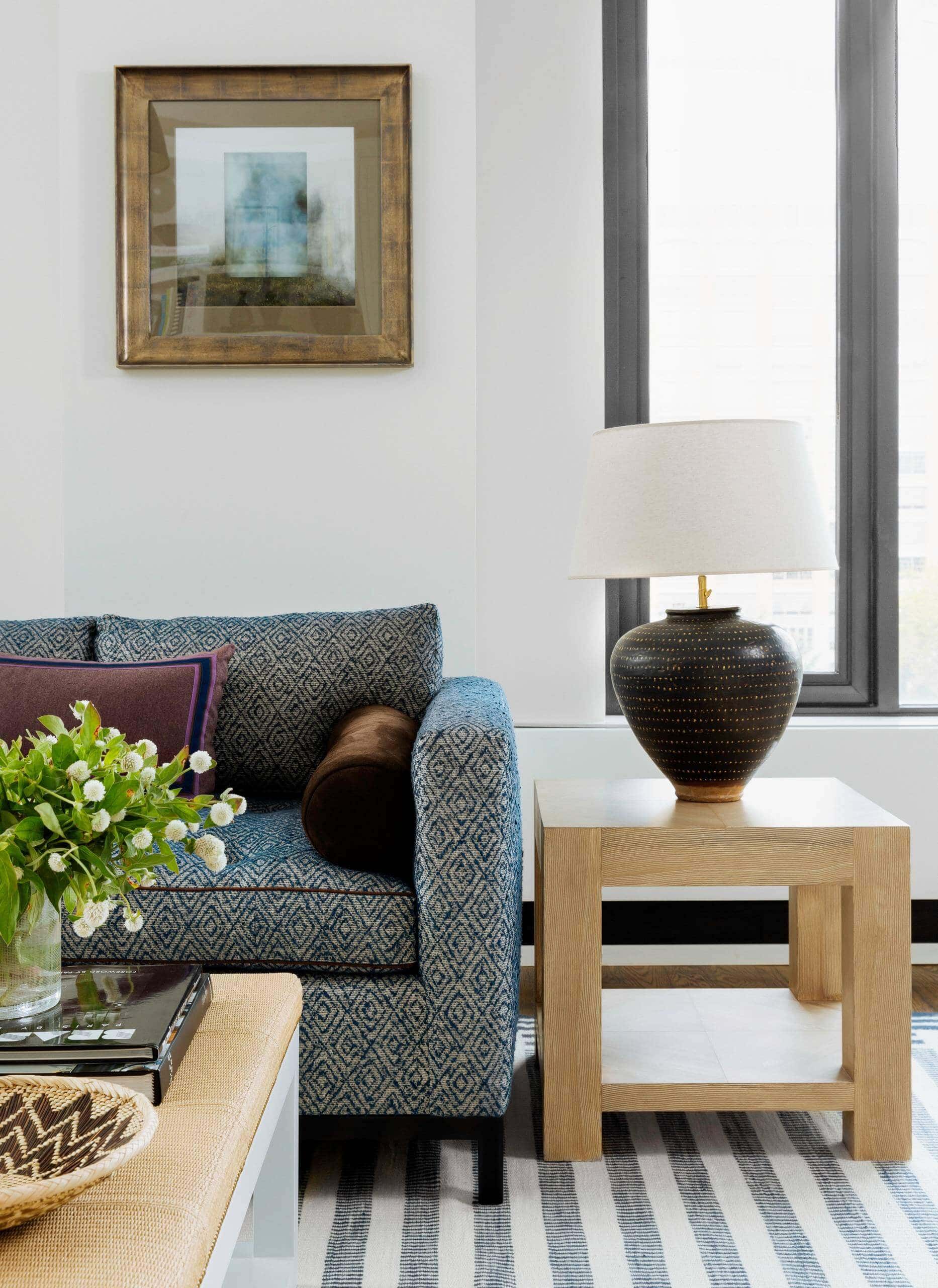 Tribeca home tour! Living room with white walls and framed artwork. Blue sofa as a focal point, accented by an artisan-inspired ceramic lamp. Transitional aesthetic with sophisticated ambiance.