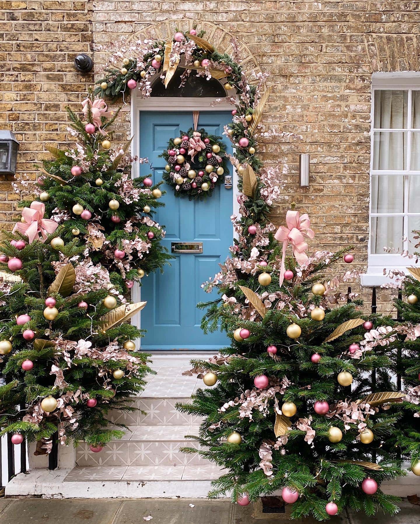 Create magical Christmas porch decor with these ideas example featuring a blue front door with Christmas trees in pink and gold porch