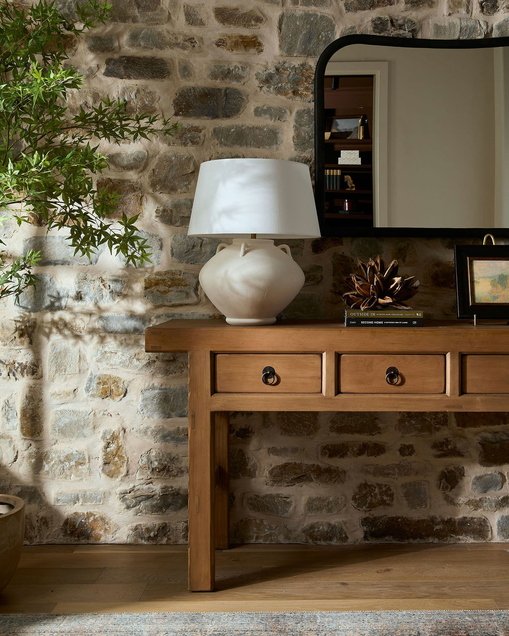 Mountain home tour entry detail with a rustic wood console against a stone wall, a white pottery lamp, and an arched black mirror.