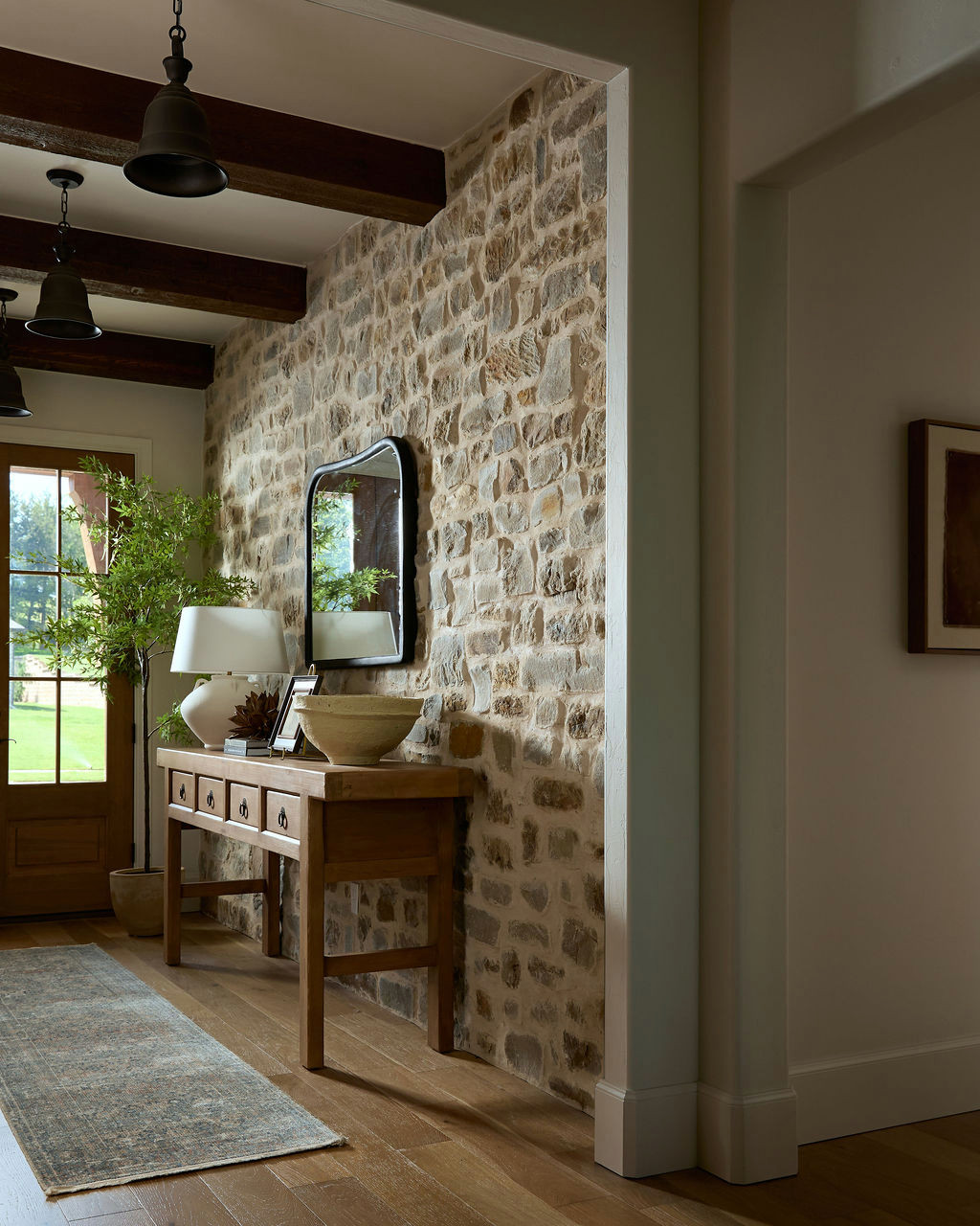 Mountain home tour foyer with a natural stone wall, dark wood beams, black bell pendants, and a wood front door.