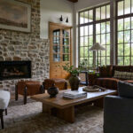 Mountain home tour living room with a stone fireplace, red plaid sofa, leather ottomans, and tall grid windows.