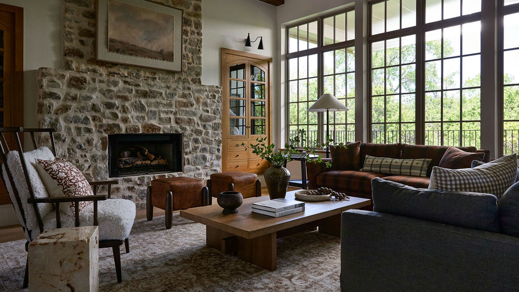 Mountain home tour living room with a stone fireplace, red plaid sofa, leather ottomans, and tall grid windows.