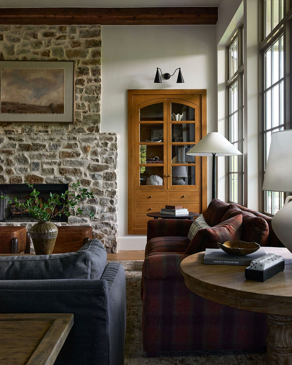 Mountain home tour living room with a stone fireplace, red plaid sofa, leather ottomans, and tall grid windows.